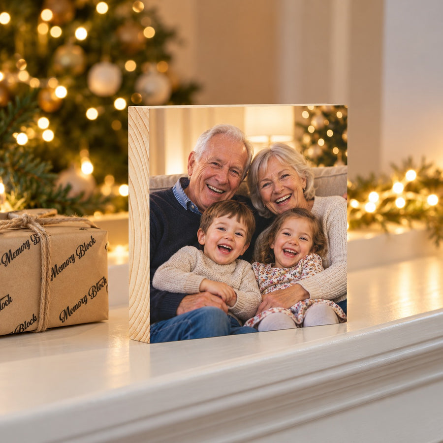 Square pine photo block of grandparents and grandchildren on a mantel, with a lit Christmas tree and a MemoryBlock-wrapped parcel