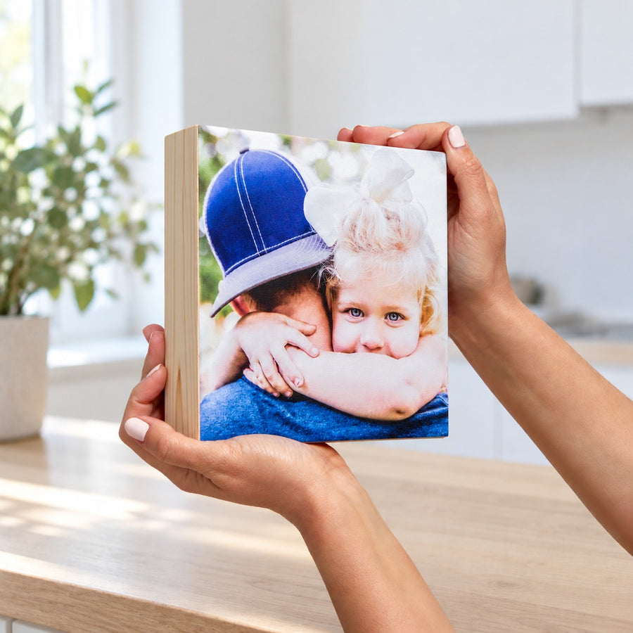Hands holding a small square pine photo block with a father and daughter hug, in a bright kitchen